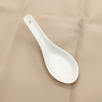 A white ceramic Chinese soup spoon with a deep, flat bowl and a short, thick handle. It sits on top of a tan tablecloth.