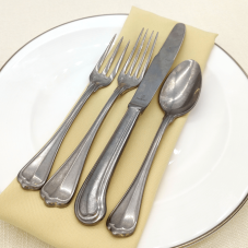 San Marco Antiqued flatware on a place setting with a yellow napkin, white plate, and pale yellow tablecloth.