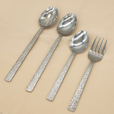 Hammered serving utensils on a tan tablecloth. From left to right, there are three serving spoons and one serving fork.
