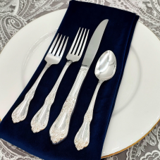 Vanessa Silver flatware on a place setting with a navy napkin, white plate, and silver lace tablecloth.