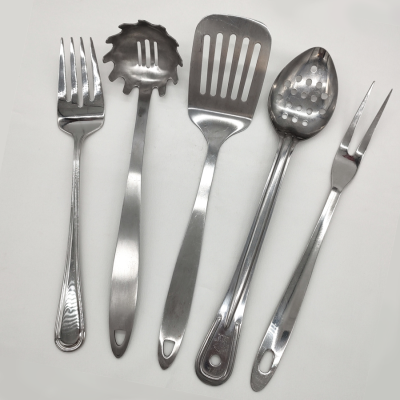 Stainless serving utensils on a white tablecloth. From left to right, there is a serving fork, a spaghetti spoon, a spatula, a slotted serving spoon, and a carving fork.