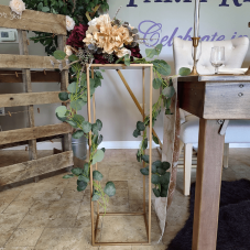 A gold, rectangular stand made of thin pieces of metal. A plant rests on top of it.