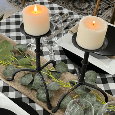 Two wrought iron candle holders with two lit candles. They sit above a burlap runner with decorative vines.